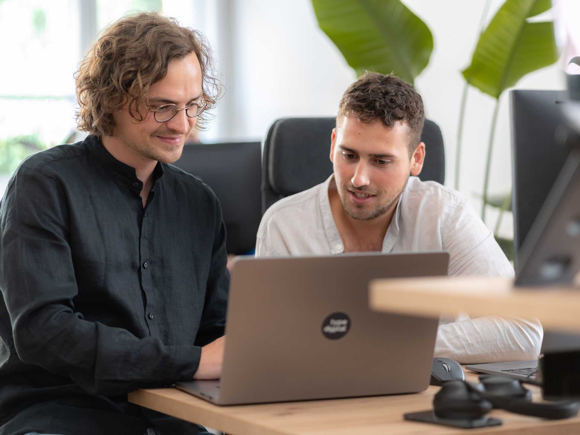 Zwei Männer sitzen nebeneinander am Tisch und blicken gemeinsam in den Laptop.