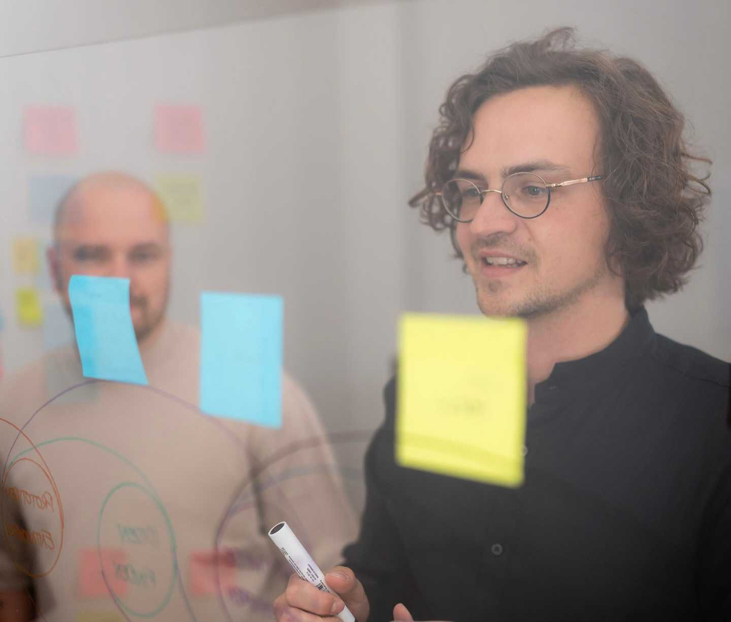 Zwei Mitarbeiter stehen vor einer Glasscheibe, an der Post its befestigt sind.