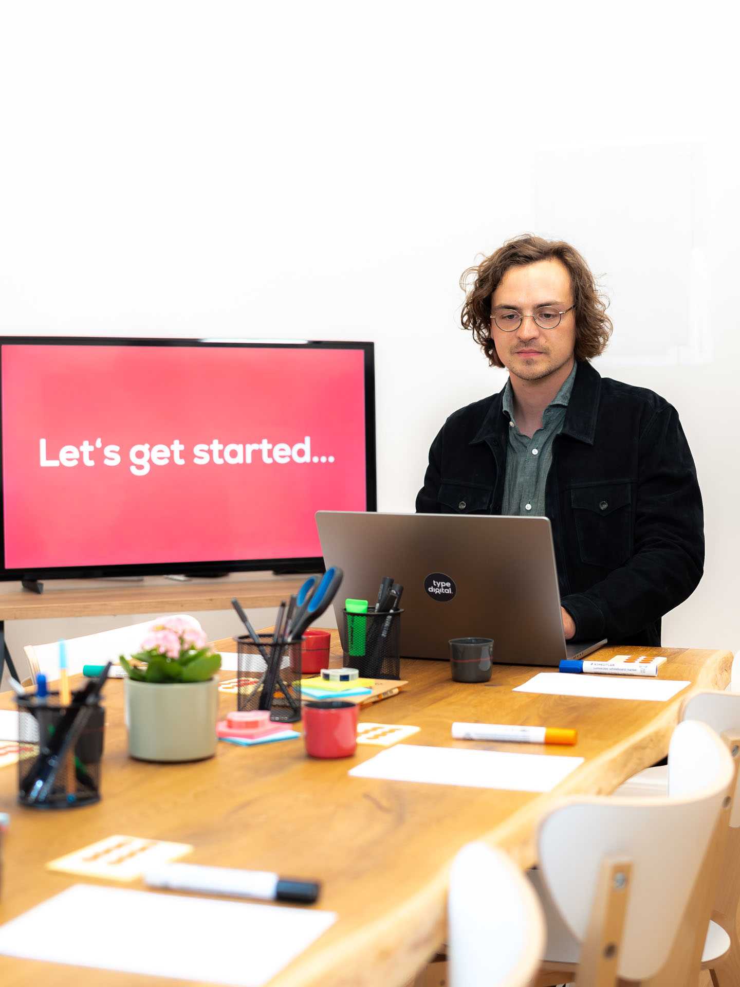 Ein Mann sitzt am Tischende und blickt in seinen Laptop vor sich. Auf dem Tisch befinden sich Utensilien für einen Workshop. Im Hintergrund befindet sich ein Fernseher auf dem der Satz "Let´s get started..." zu lesen ist.